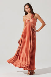 A woman is wearing a floor-length, pleated, one-shoulder gown in a warm, coral-orange shade. The dress features a twisted strap detail at the neckline and a small cutout beneath the bust. The pleats run vertically from the waist to the hem, creating a flowing silhouette. She is wearing open-toed heels that complement the dress.